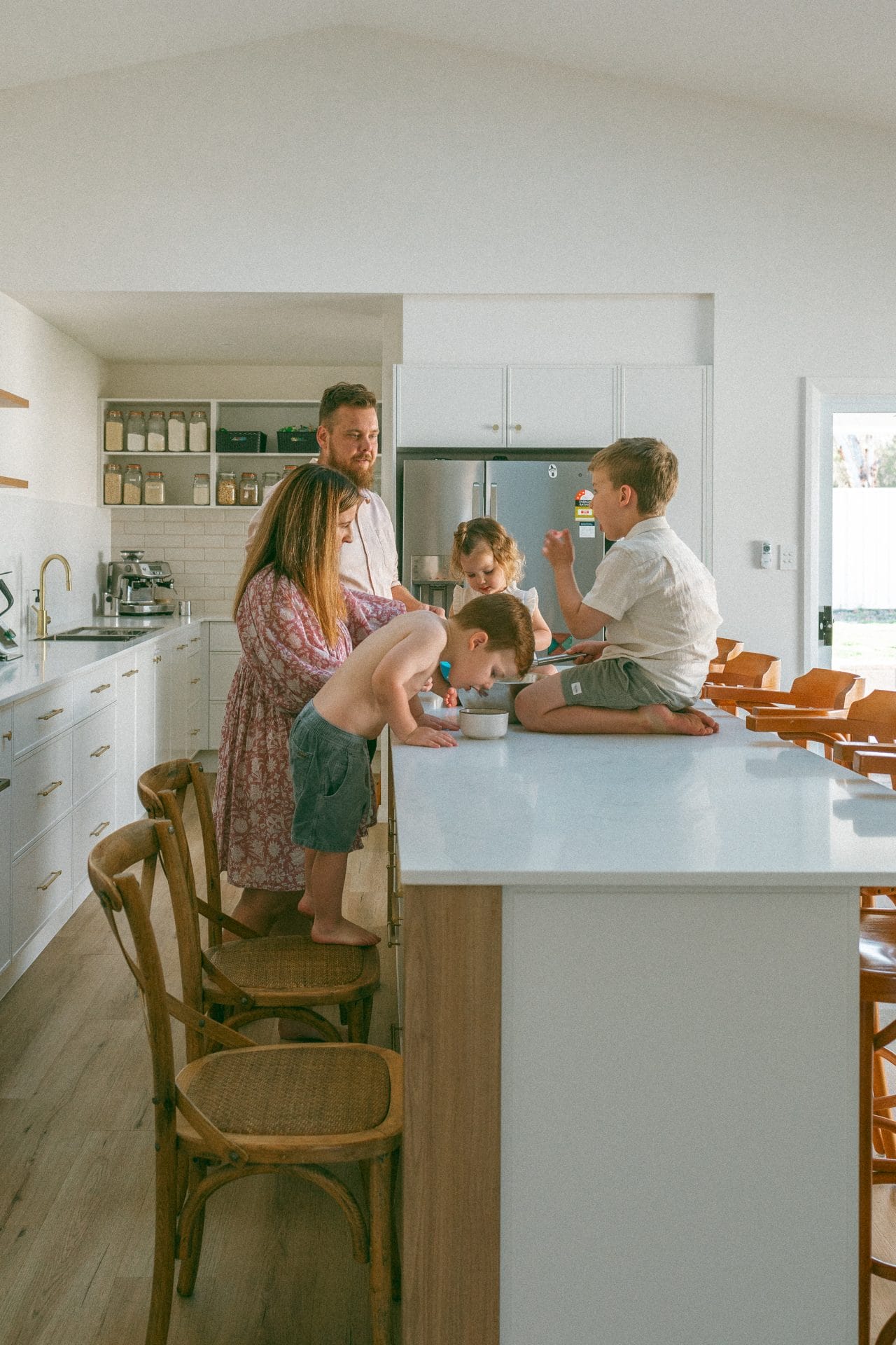 A family of five enjoying time together in a bright kitchen, with two adults smiling at three children who are sitting at a counter.
