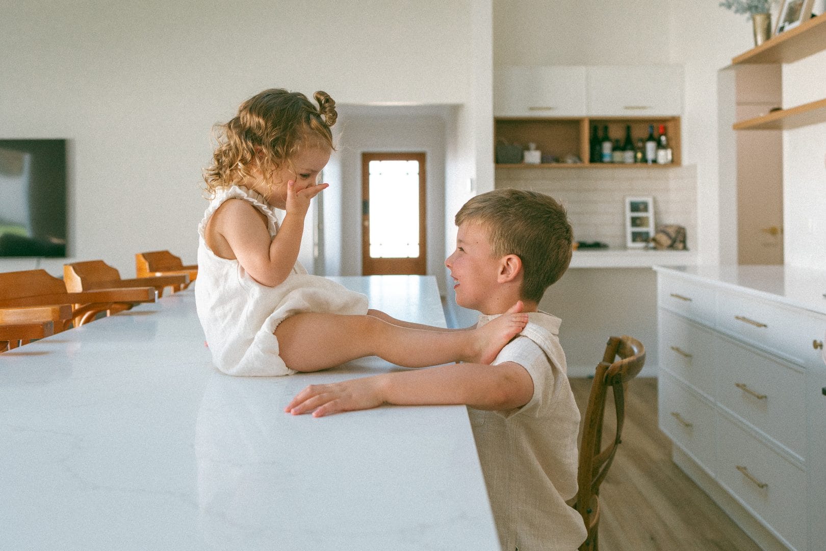 A young boy and a toddler girl sitting at a kitchen counter, engaging in a playful conversation in a bright, modern kitchen.