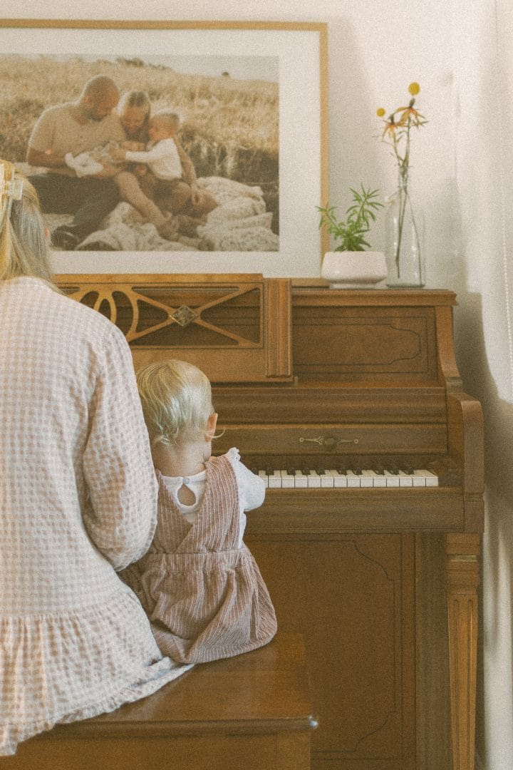 A toddler in a pink dress sits at an old piano, looking at a framed family photo, with a woman's back visible in the foreground.