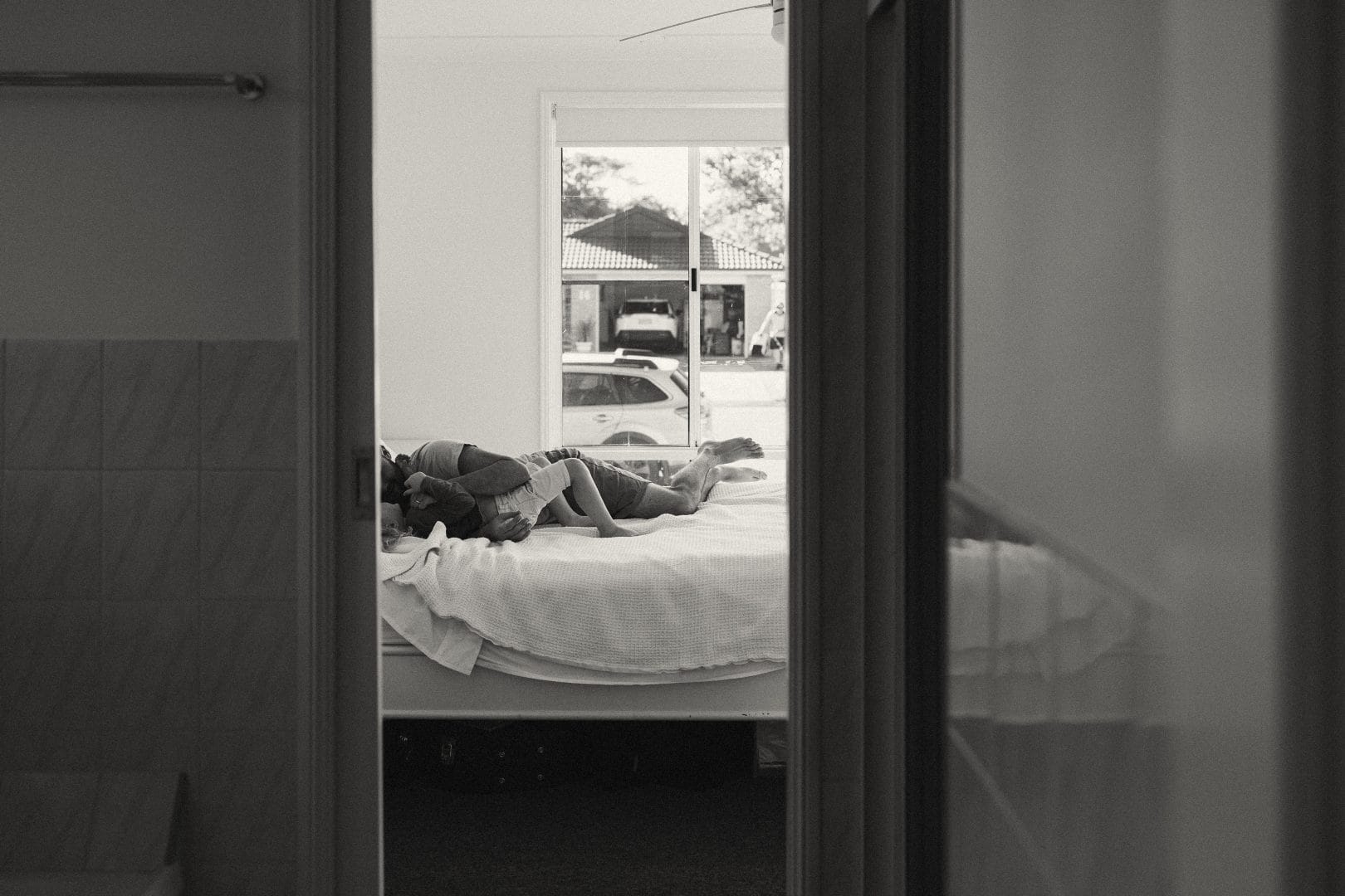 Black and white photo of a person lying on a bed viewed from an open doorway, with a glimpse of a street scene visible through another door across the room.