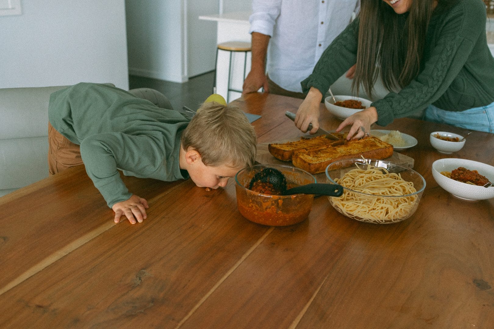 A young boy leans over a dining table to smell food as adults prepare plates around him. the table is laden with dishes like spaghetti and bread.
