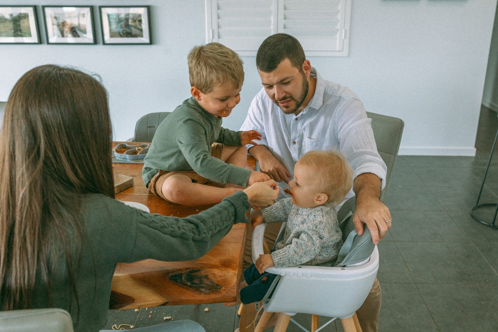 A family with two young children gathered around a dining table, interacting affectionately with the father assisting the toddler while the mother watches.