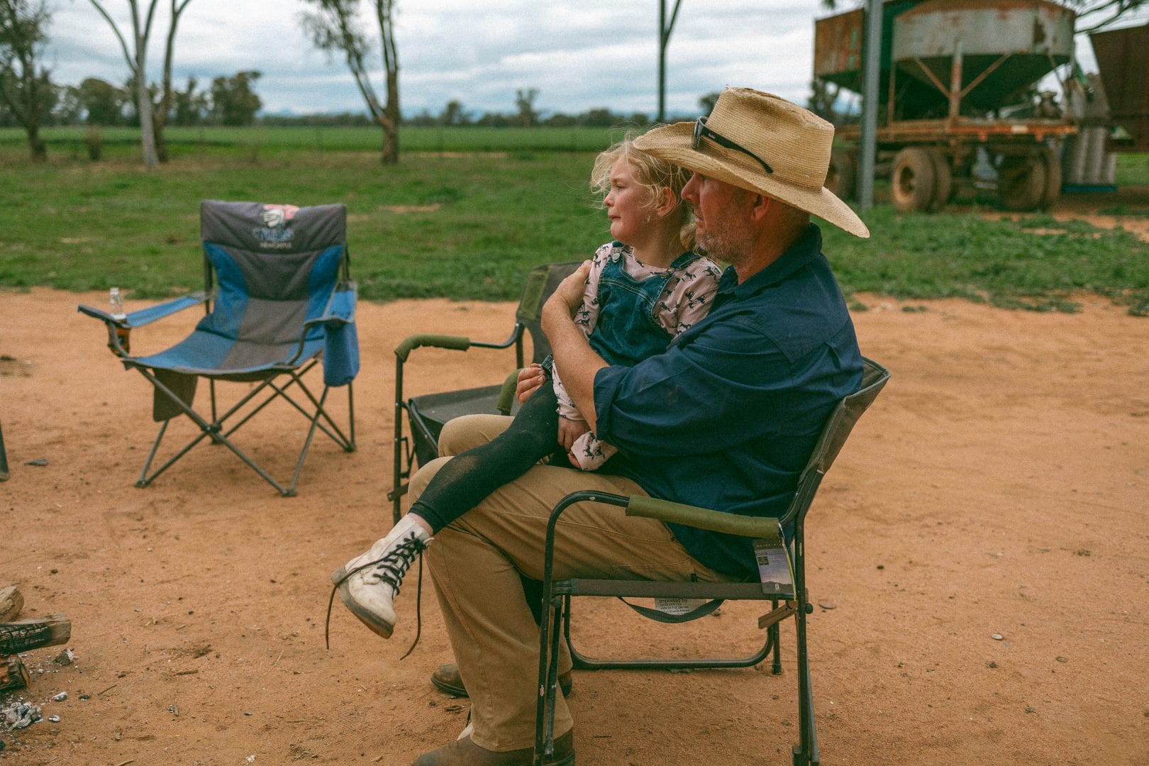 An older man and a young girl sit on folding chairs in a rural area, both wearing hats and looking into the distance.