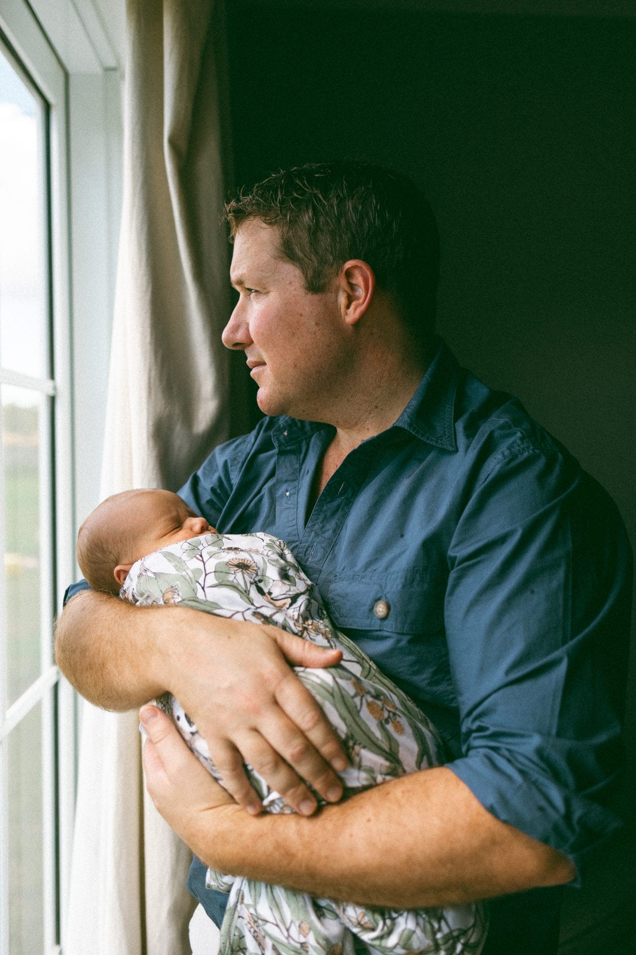A man in a blue shirt holds a sleeping baby by a window, looking thoughtfully outside.