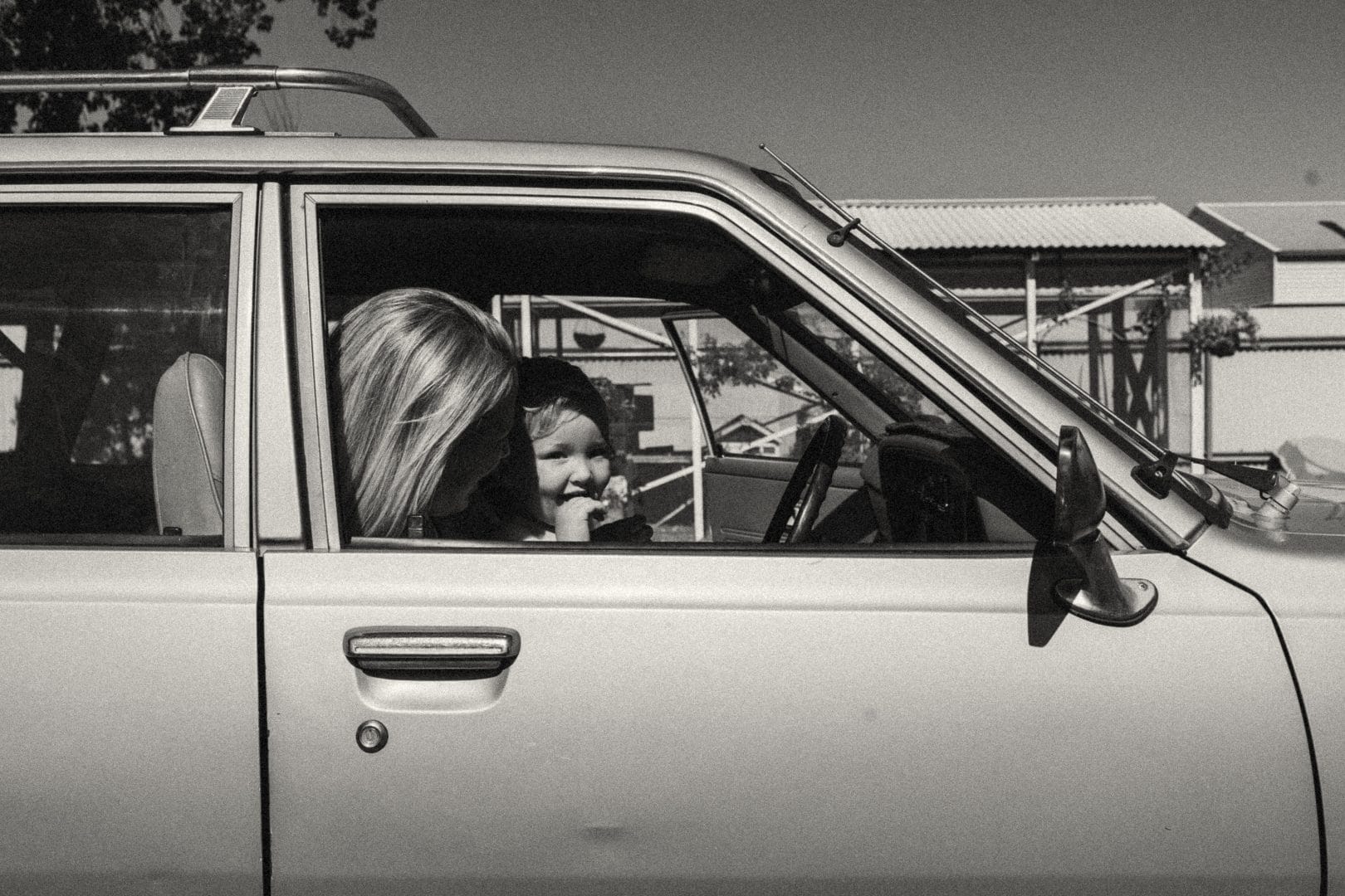 Black and white photo of a smiling child peeking through the rear window of a car, with a woman in the driver's seat.