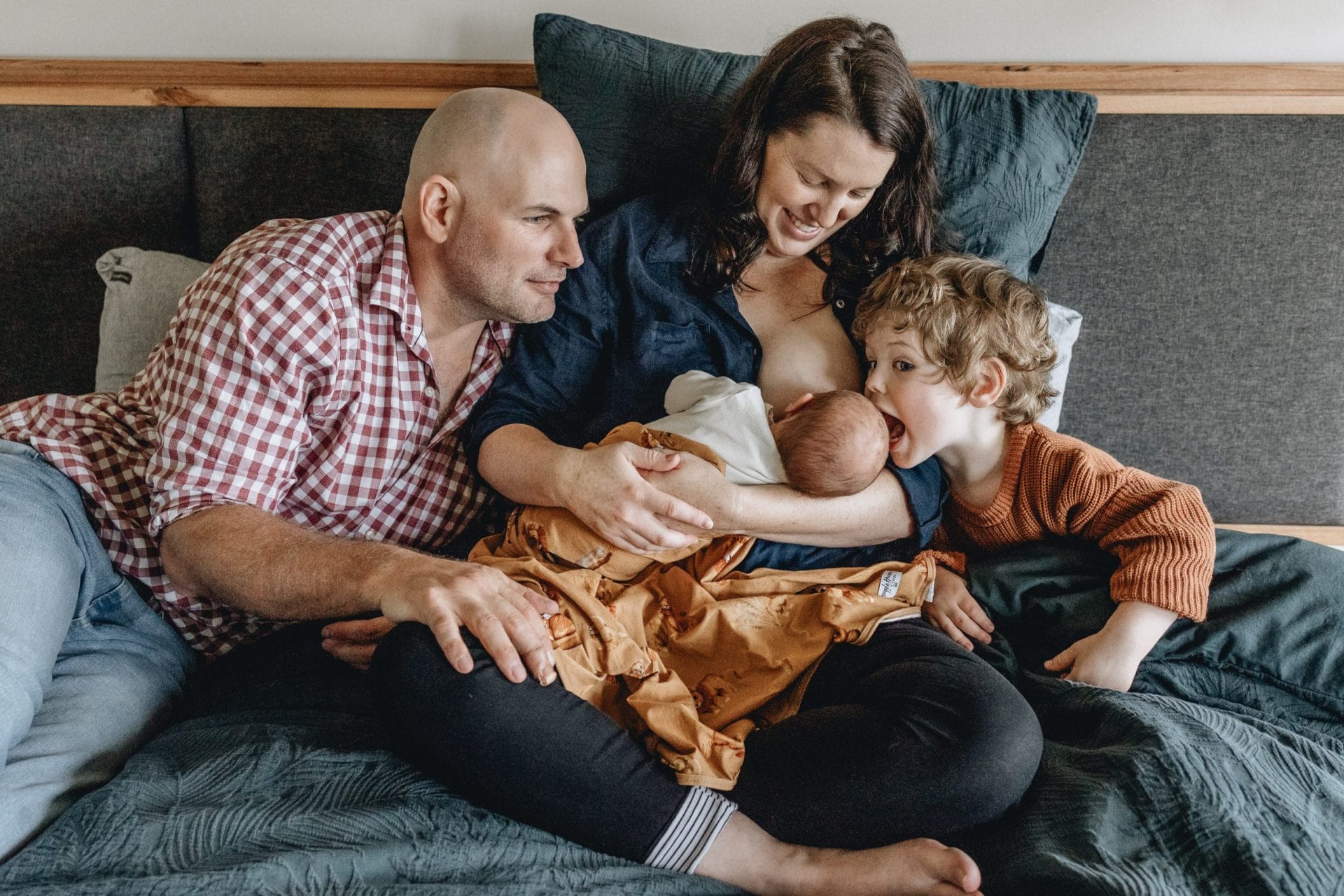 A family of four with two young children sitting together on a bed, sharing a joyful moment.