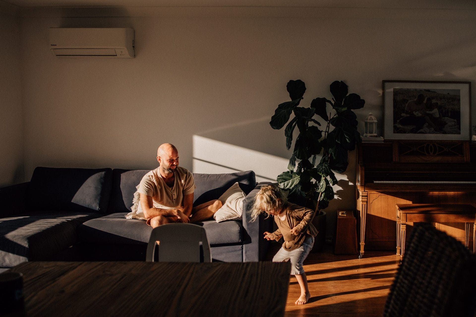 A man and a young child play in a sunlit living room with a blue sofa, wooden furniture, and a large plant.