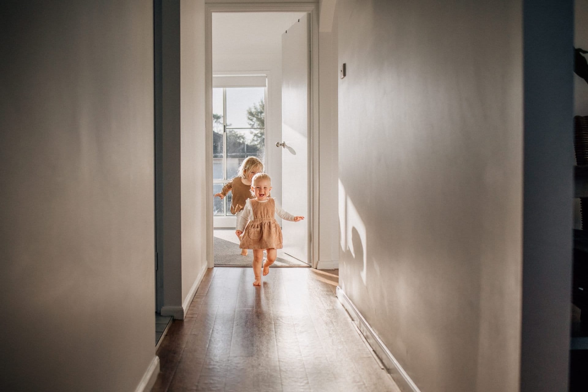 Two young children run joyfully down a sunlit hallway toward an open door.