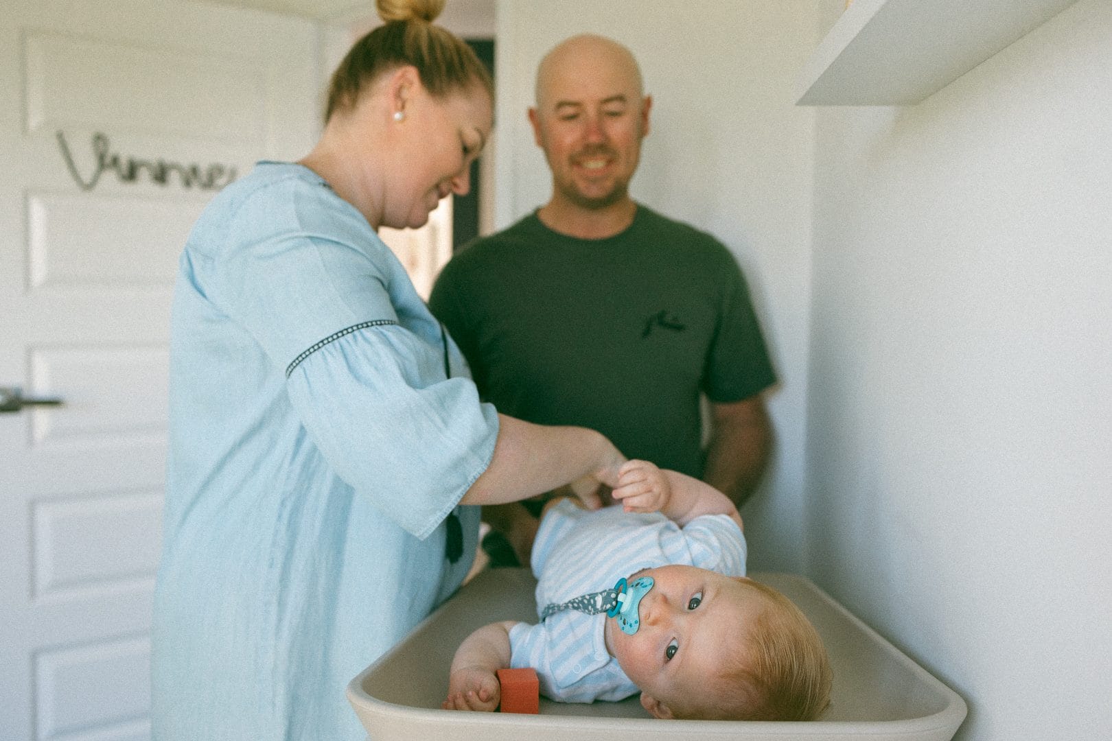 A woman and a man smiling at a baby lying in a changing table, in a bright, cozy room.