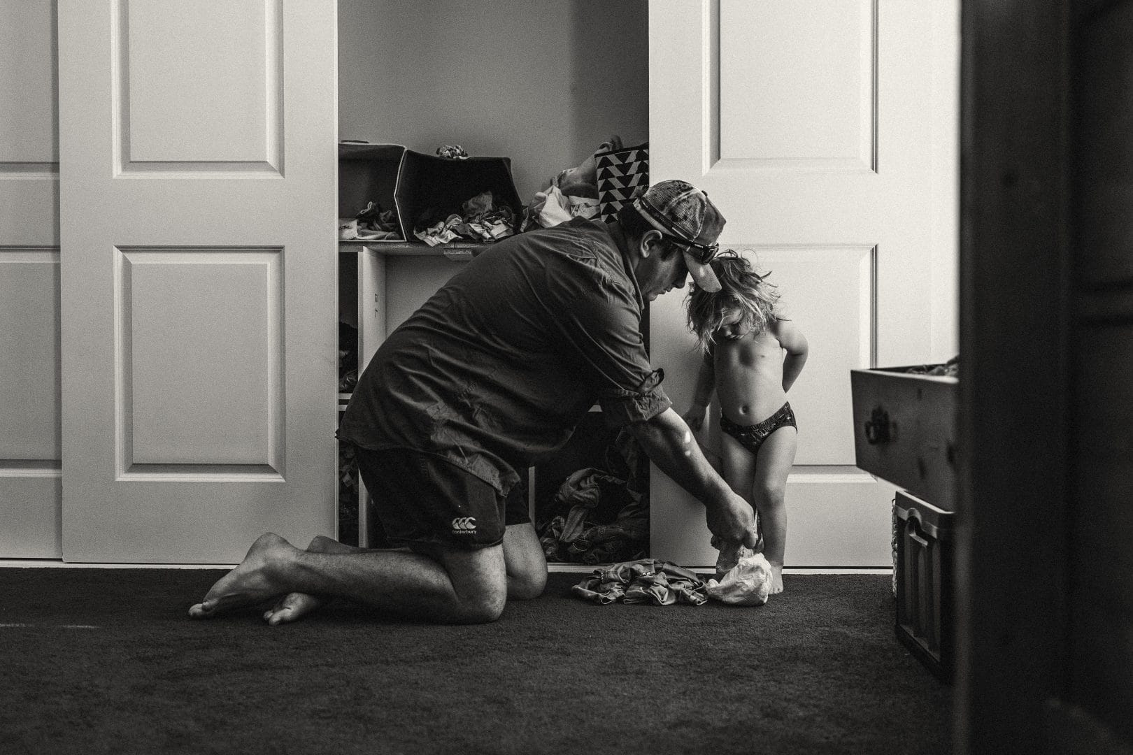 A man kneeling assists a young child in a diaper with dressing in a cluttered room, in a black and white photo.